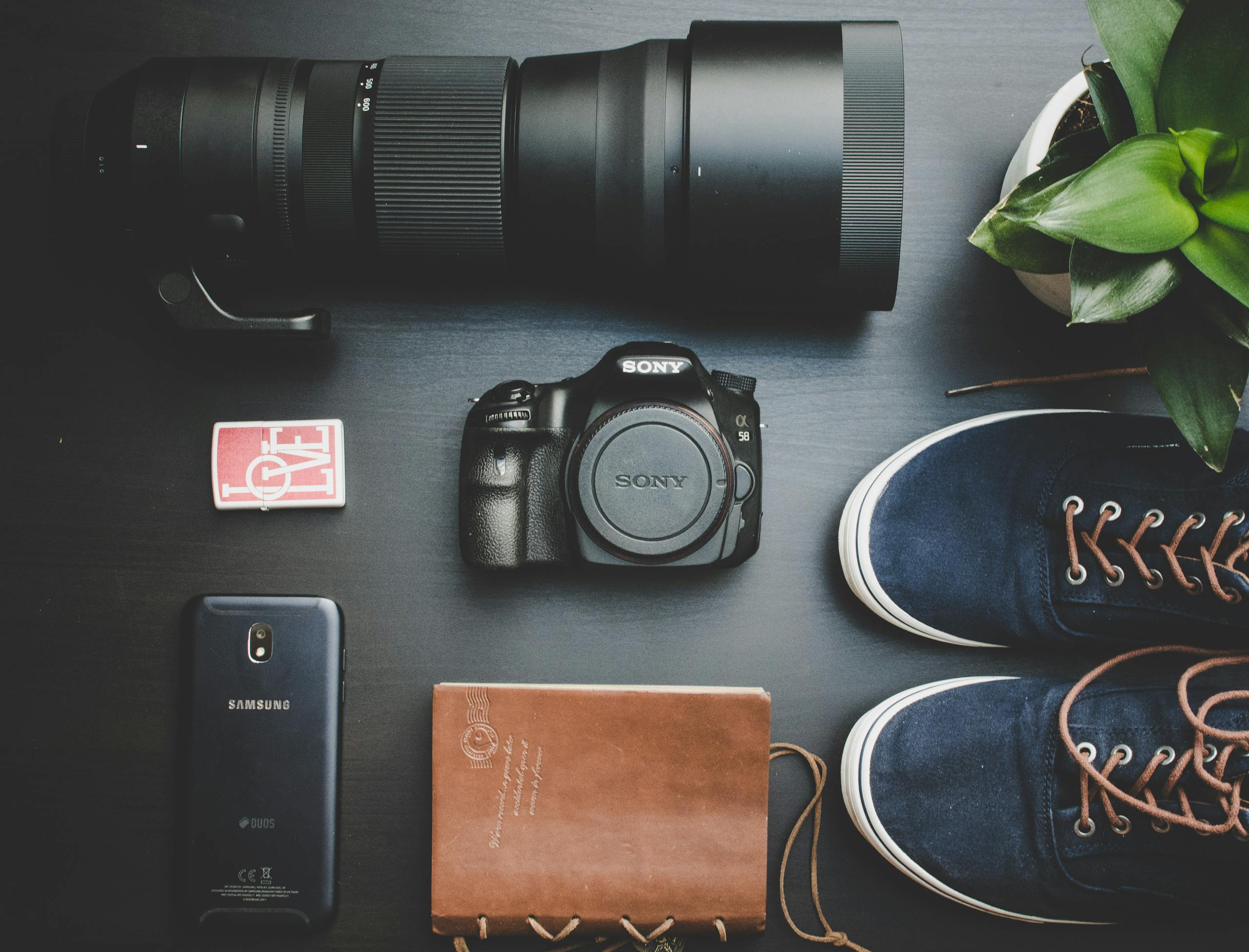 Creative flat lay of camera equipment, smartphone, notebook, shoes, and plant on a wooden surface.